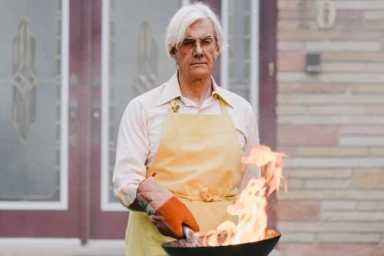 Elderly man with white hair wearing a yellow apron and an oven mitt, holding a pan with visible flames
