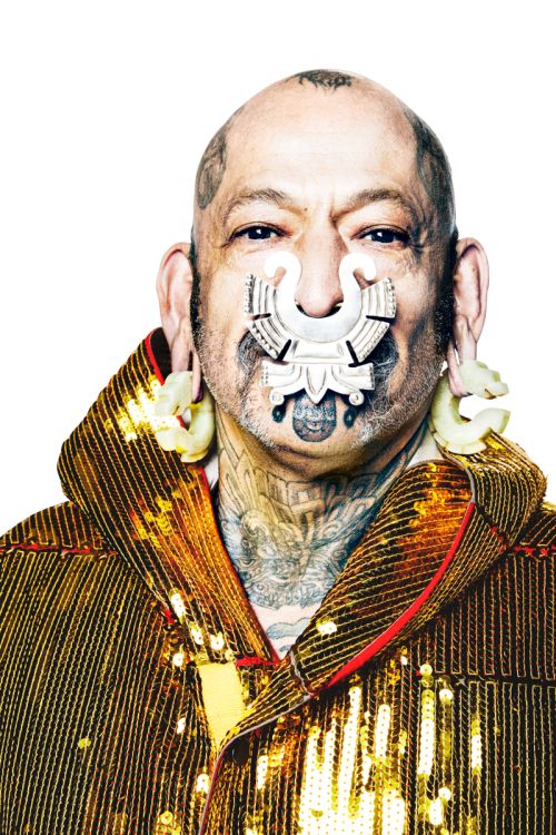 Portrait of an adult male with visible tattoos and face jewelry wearing a gold sequin jacket against a white background in a photo studio.
