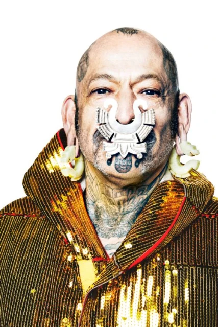 Portrait of an adult male with visible tattoos and face jewelry wearing a gold sequin jacket against a white background in a photo studio.
