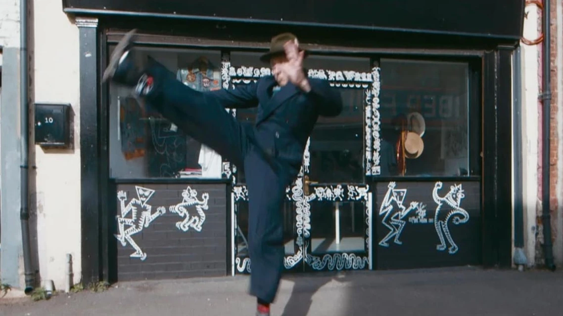 Adult in suit with hat poses with high kick outside a shop with graffiti and hats displayed in window on a sunny day.
