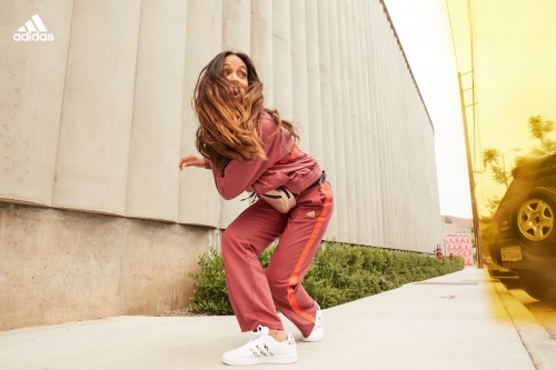 An athletic woman in a maroon track suit strikes a dynamic pose on a city sidewalk, with an Adidas logo visible in the upper-left corner.