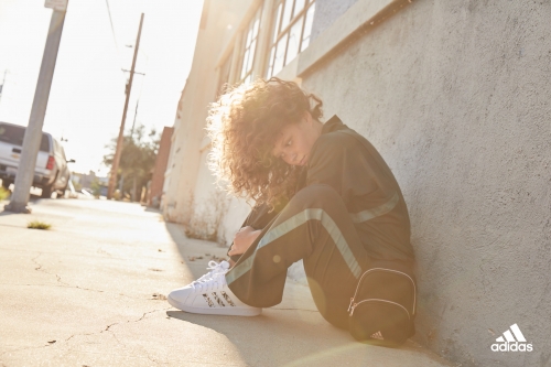 A young woman with curly hair wearing a dark green tracksuit and white sneakers poses against a wall outdoors on a sunny day.