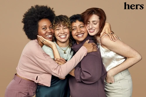 Four smiling women of diverse ethnicities embrace indoors against a neutral backdrop in a beauty and lifestyle shot with soft lighting.