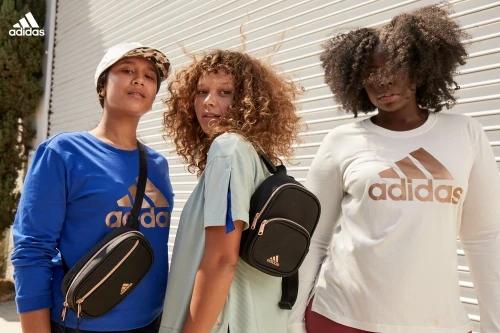 Three women wearing Adidas apparel and accessories pose outdoors against a white garage door, showcasing fashion and active lifestyle.