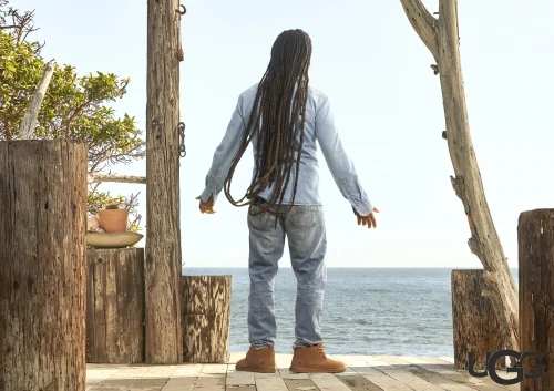 A man stands on a wooden platform facing the ocean, wearing a light blue shirt, jeans, and brown UGG boots, with dreadlocks hanging down his back.