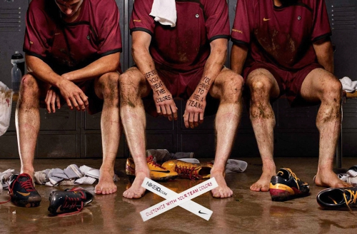Three mud-covered soccer players sit in a locker room; they wear maroon jerseys, shorts, and mud. Soccer shoes and a water bottle are visible.