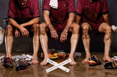 Three mud-covered soccer players sit in a locker room; they wear maroon jerseys, shorts, and mud. Soccer shoes and a water bottle are visible.