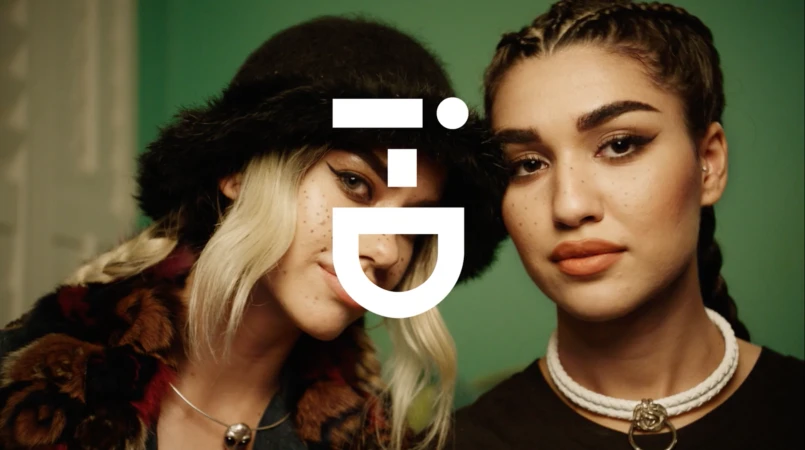 Close-up portrait of two young women with styled hair and makeup standing together indoors against a green wall