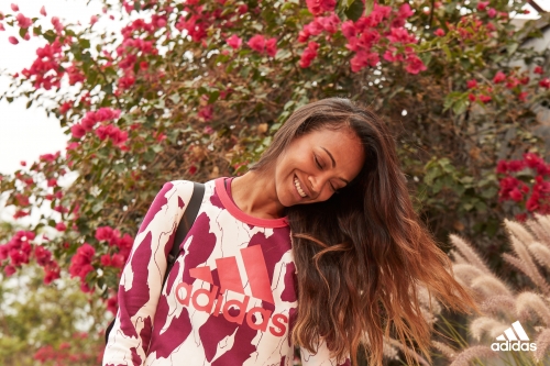 Smiling woman with long brown hair in a printed Adidas sweatshirt outdoors among pink bougainvillea and tall grasses.