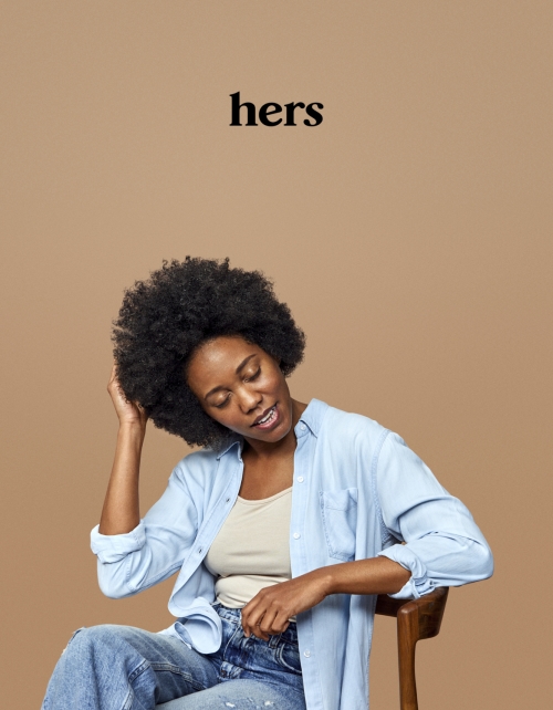Adult woman with afro hairstyle sits in chair wearing denim in a neutral studio. She touches her hair while wearing an unbuttoned light-blue shirt.