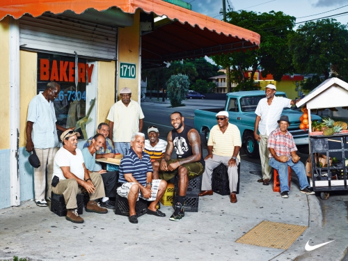 Group of senior men gather outdoors near a bakery and a fruit cart with Lebron James sitting among them in casual clothes, vintage truck parked behind.
