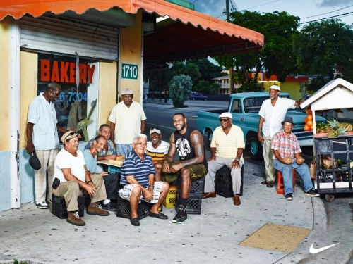Group of senior men gather outdoors near a bakery and a fruit cart with Lebron James sitting among them in casual clothes, vintage truck parked behind.