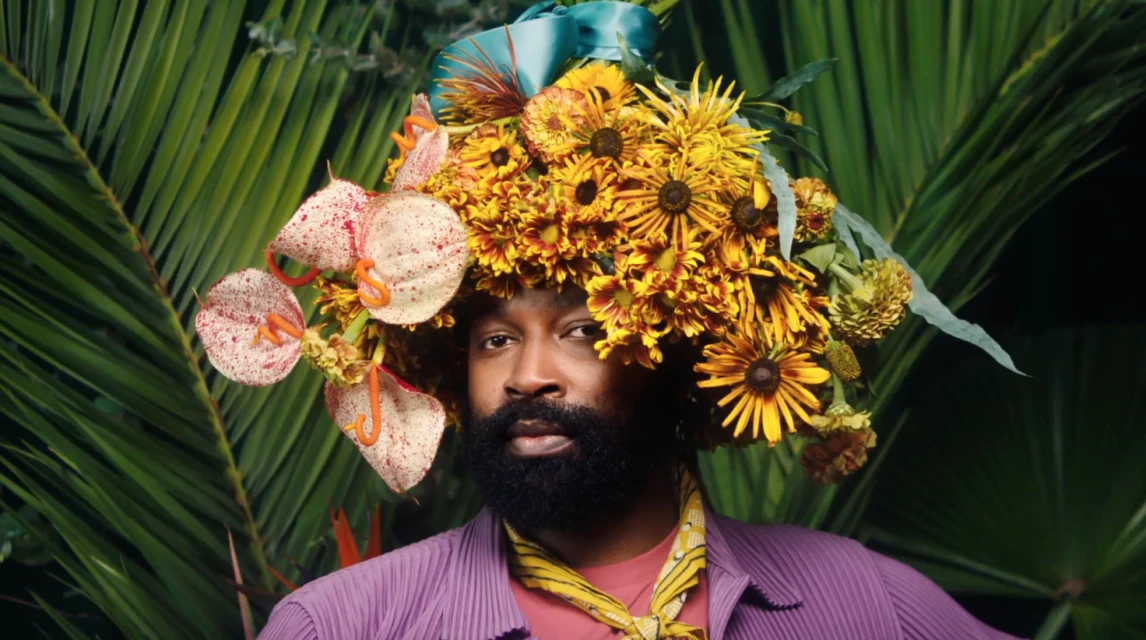 Adult male portrait with a beard, wearing a purple shirt and flowers on his head, set against a lush green backdrop.
