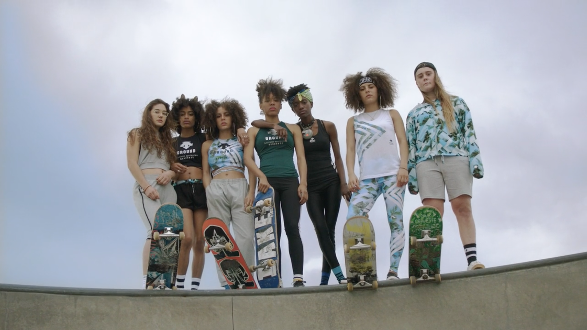 Group of diverse young women skaters in sportswear and casual wear stand together at a concrete skatepark, holding skateboards.
