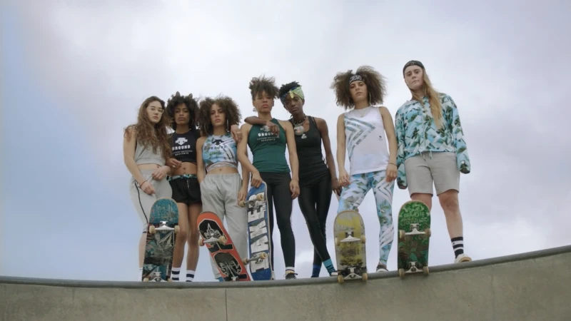 Group of diverse young women skaters in sportswear and casual wear stand together at a concrete skatepark, holding skateboards.