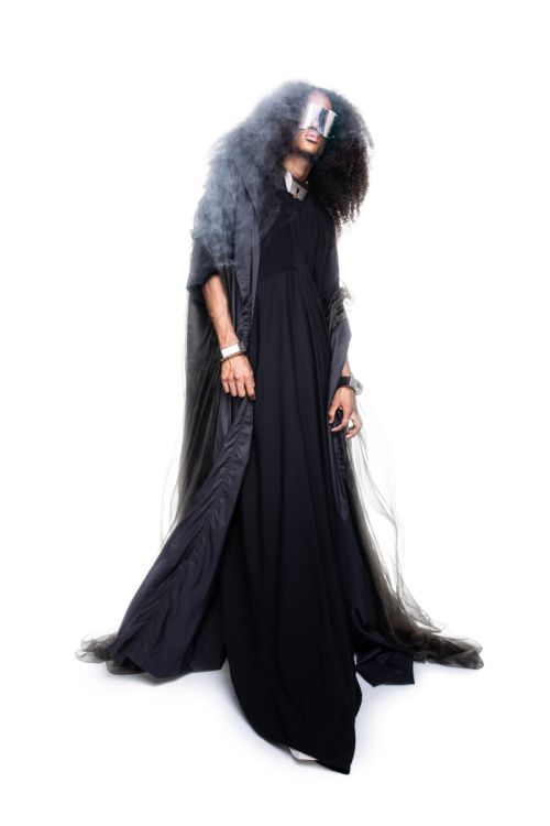 Model in a long black dress and hooded dark grey cloak wearing reflective visor glasses against a bright white background