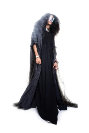 Model in a long black dress and hooded dark grey cloak wearing reflective visor glasses against a bright white background