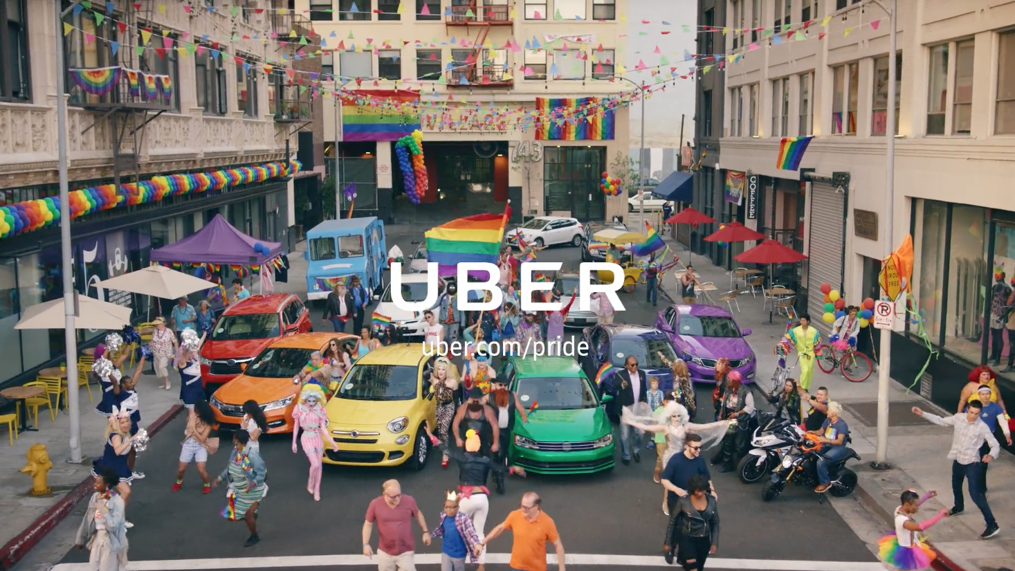 Wide shot of a street parade with diverse people, rainbow flags, and colorful decorated vehicles, promoting Uber during a Pride event.