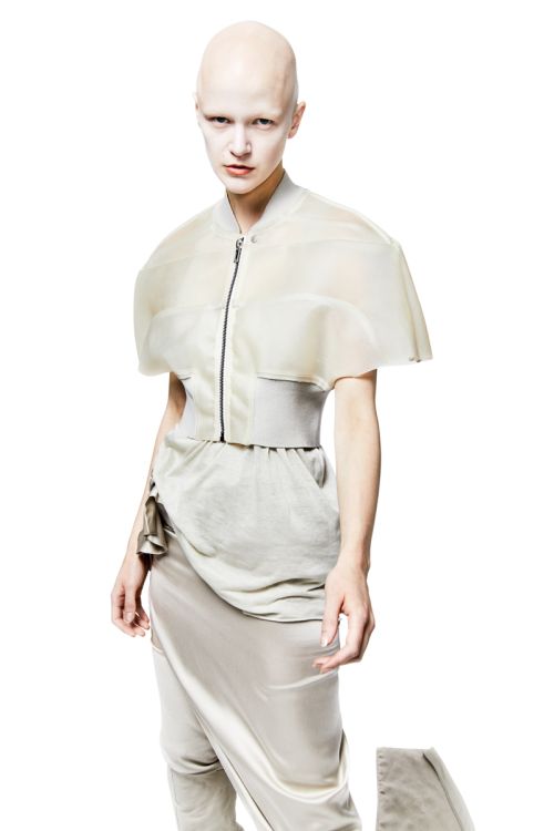 A pale, bald person poses in a studio wearing a sheer cape-like jacket, high-waisted belted pants, and draped skirt against a white backdrop.