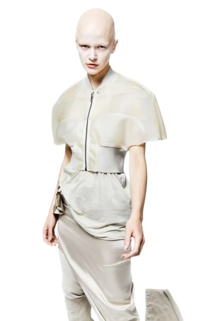 A pale, bald person poses in a studio wearing a sheer cape-like jacket, high-waisted belted pants, and draped skirt against a white backdrop.