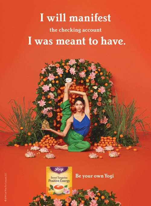 Adult female in yoga pose surrounded by flowers and tangerines, advertising Yogi Sweet Tangerine Positive Energy tea against orange backdrop.