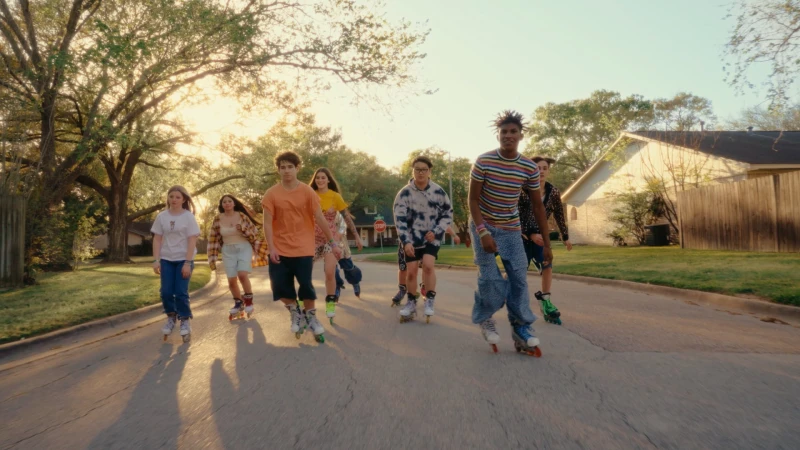 A group of seven teenagers roller skating down a suburban street lined with houses and mature trees during the daytime with natural sunlight.