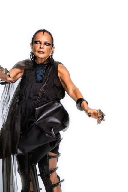 Woman poses in dramatic black outfit with sheer cape, bold eye makeup, cuff bracelet and ring on a white background in a studio shot.