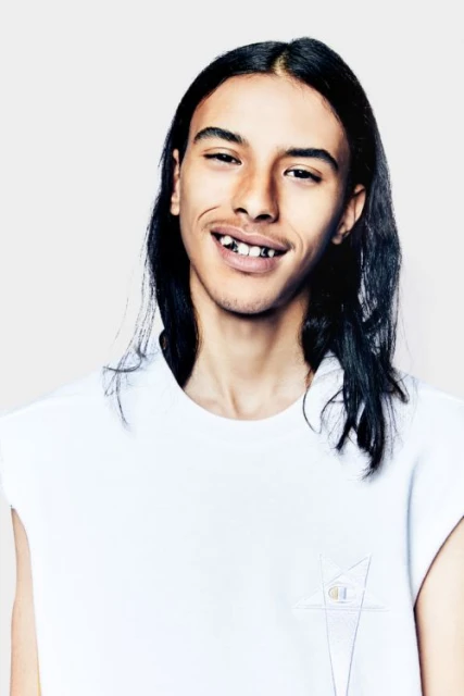 Young man with long hair smiles in a fashion-style portrait with a white sleeveless shirt in front of a solid grey background.