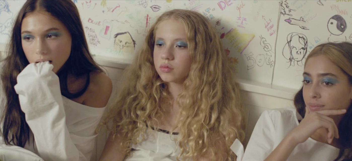 Three young women with bright blue eyeshadow pose in front of a scribbled wall, wearing white tops in a close-up portrait.