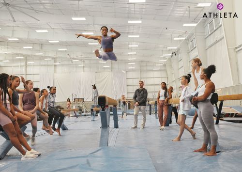 Female gymnast in mid-air leap during floor exercise, observed by a group of young athletes, in a brightly lit gymnasium.