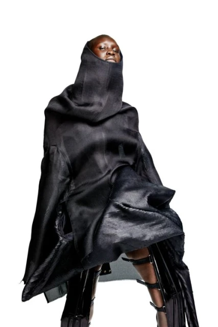 Fashion model wearing a dramatic black hooded costume with shin-high leather boots, set against a bright white backdrop in a studio shot.