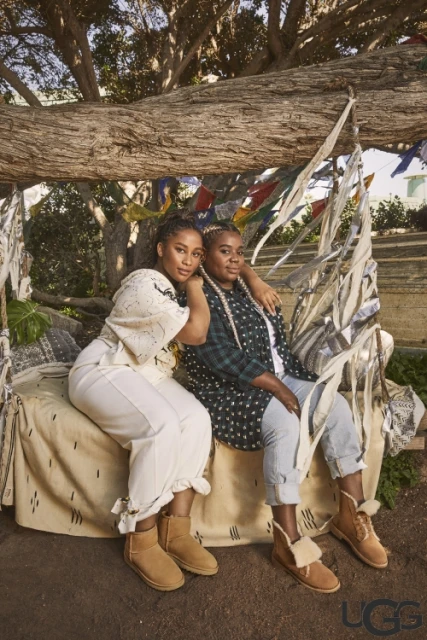 Two adult females sit on a patterned tan blanket under a tree, wearing casual clothing and UGG brand sheepskin boots in outdoor setting.