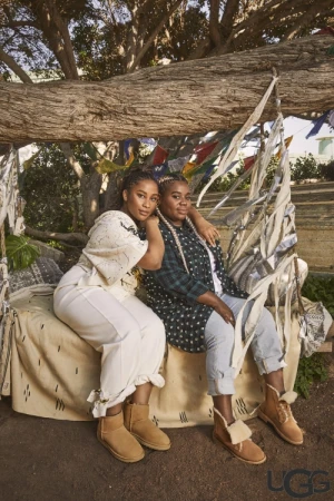 Two adult females sit on a patterned tan blanket under a tree, wearing casual clothing and UGG brand sheepskin boots in outdoor setting.