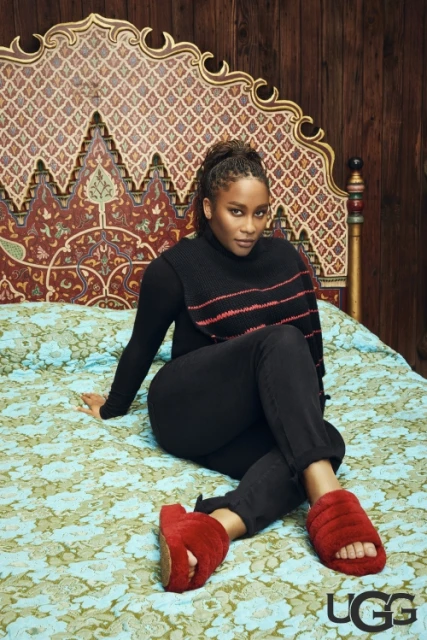 Woman in black sweater and pants sits on a bed with a patterned headboard and floral bedding, wearing UGG slippers and posing indoors.