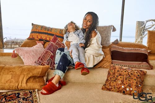Smiling Black mother and child, sitting on a couch with decorative pillows near a window overlooking the ocean, wearing UGG slippers.
