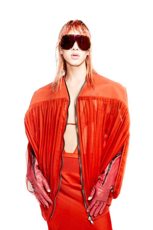 Person with orange hair wearing an orange bomber jacket, skirt, and sunglasses against a white backdrop in a studio shot.