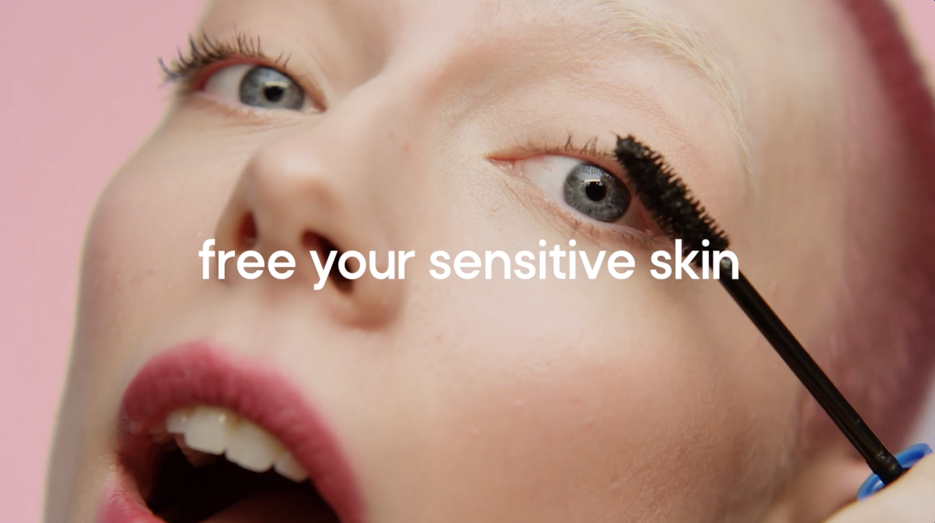 Close-up of fair-skinned woman applying black mascara with pink background and text overlay, free your sensitive skin.