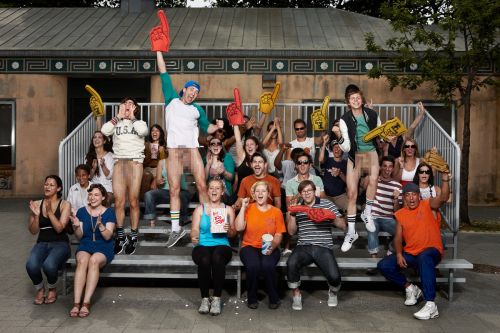 A crowd of partially nude people on bleachers, wearing sports fan apparel such as foam fingers and hats, outdoors in daylight.