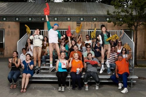A crowd of partially nude people on bleachers, wearing sports fan apparel such as foam fingers and hats, outdoors in daylight.