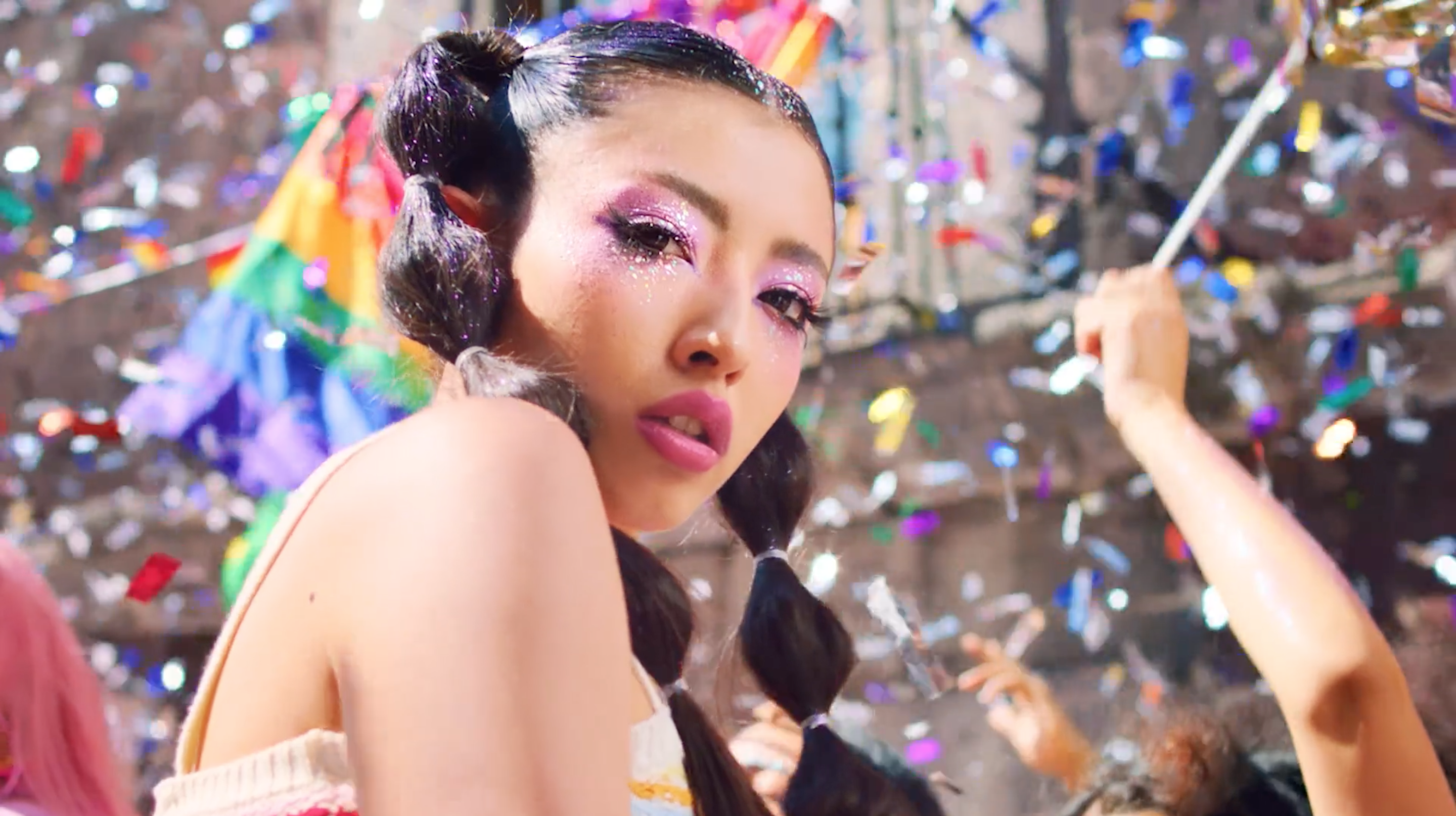 Close-up portrait of a young woman with pink makeup, pigtails, and colorful confetti and pride flag in the background at a celebration.