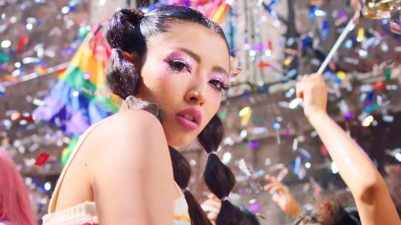 Close-up portrait of a young woman with pink makeup, pigtails, and colorful confetti and pride flag in the background at a celebration.