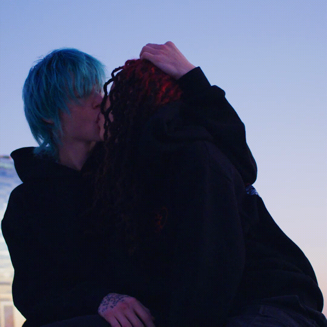 A close-up captures a couple embracing outdoors with a gradient blue sky backdrop, showing blue and red dyed hair and dark hooded sweatshirts.