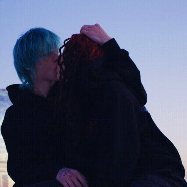 A close-up captures a couple embracing outdoors with a gradient blue sky backdrop, showing blue and red dyed hair and dark hooded sweatshirts.