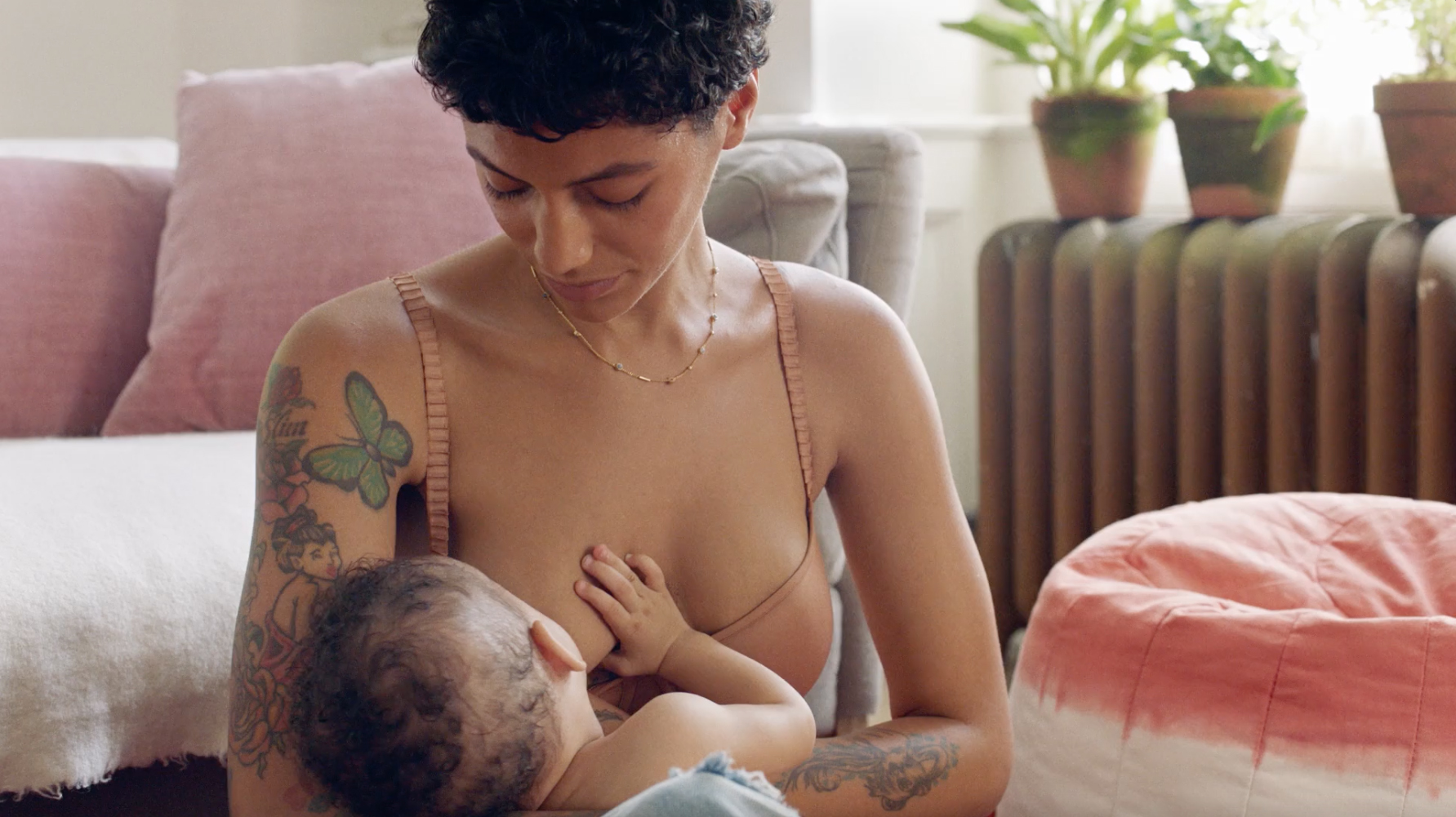 Adult woman breastfeeding a baby indoors near a radiator and houseplants, with sunlight streaming through the window in a home setting.