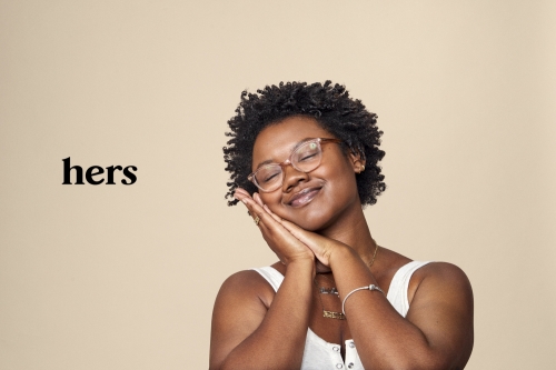 Portrait of a smiling African-American woman with eyeglasses, hands near her face, tank top, against a neutral backdrop with 'hers' text.