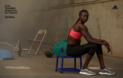 Athletic woman in workout clothes poses on a blue stool indoors. Weights and other exercise equipment are visible in the background.