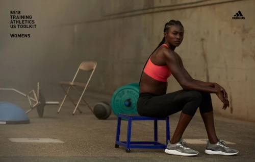 Athletic woman in workout clothes poses on a blue stool indoors. Weights and other exercise equipment are visible in the background.