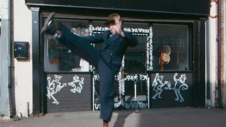 Man wearing a dark suit and hat kicking in the air outside a storefront with white chalk illustrations on a dark brick exterior.