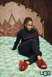 Black woman wearing a black turtleneck and red fluffy slippers seated on a floral bed with an ornate headboard