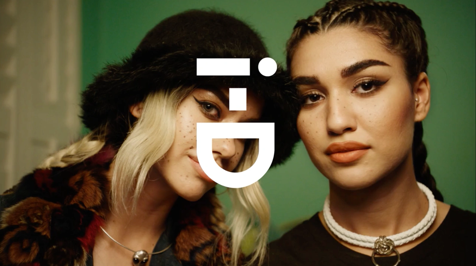 Close-up portrait of two young women with styled hair and makeup standing together indoors against a green wall